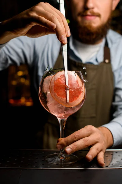 Kelchglas mit kaltem Cocktail, das die Hand des Barmannes mit einer Pinzette mit frischen Scheiben Grapefruit dekoriert — Stockfoto