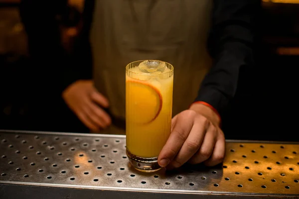 Nahaufnahme des Glases mit kaltem alkoholischen Cocktail mit Orangenscheiben ist auf der Theke und wird von männlicher Hand angefasst — Stockfoto