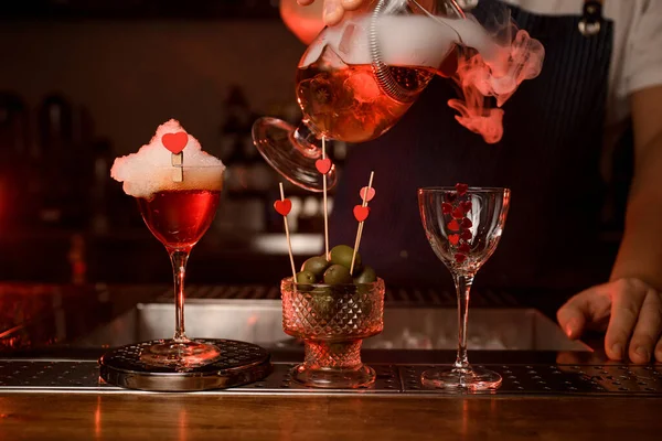 Gran vista en el vaso de cóctel con espuma y jarrón con aceitunas y vidrio en el que el camarero vierte bebidas humeantes — Foto de Stock