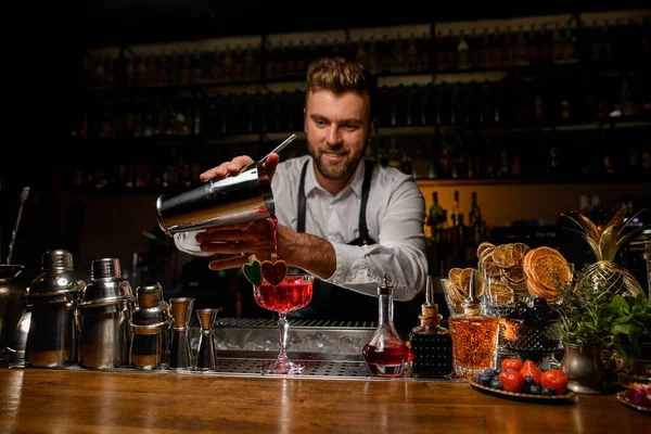 Barman sorridente ordenadamente derramando bebida alcoólica fresca vermelha em vidro — Fotografia de Stock