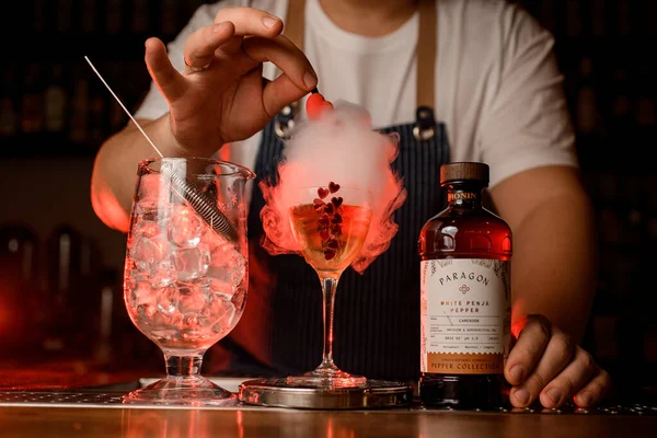 UKRAINE, KYIV - NOVEMBER 25, 2021: male hand pierced smoky air bubble in glass. Mixing cup with ice and bottle of paragon by Monin nearby on bar — стоковое фото