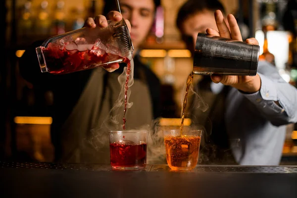 Вид двух прозрачных бокалов со льдом, в которые бармены тщательно наливают различные коктейли на пару — стоковое фото