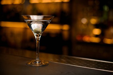 İçecekli martini bardağı ve tezgahta yeşil zeytin. Bulanık çubuk arkaplanı.