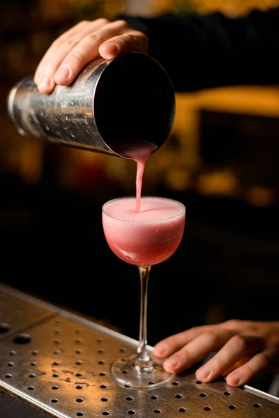 Barista professionalmente versa cocktail rosa da shaker in vetro in piedi su bancone bar in metallo — Foto Stock