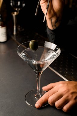 Dövmeli, cımbız tutan ve martini bardağına yeşil zeytin koyan bir el.