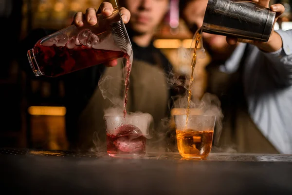 Dvě sklenice s kostkami ledu stojí na baru a dva barmani je naplní alkoholickými nápoji — Stock fotografie
