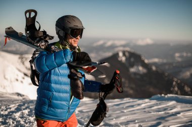 Gözlüklü, kayak kasklı ve omuzlarında kayaklarla gülümseyen bir adam.