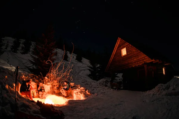 gorgeous view of wooden house and a bonfire with people around it on snow-covered mountainside at night