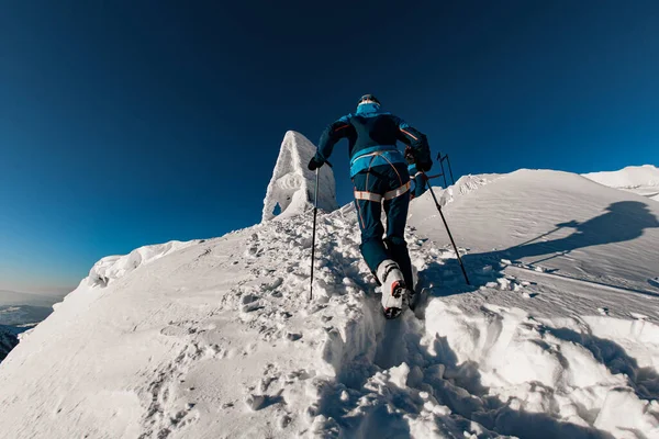 Πίσω όψη ενός άνδρα με ένα φωτεινό κοστούμι σκι με κοντάρια σκι σκαρφαλώνοντας σε ένα μονοπάτι χιονιού — Φωτογραφία Αρχείου