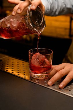 Barmenin eli süzgeci tutuyor ve bardakla içkiyi karıştırıyor ve buzlu bardağa döküyor.