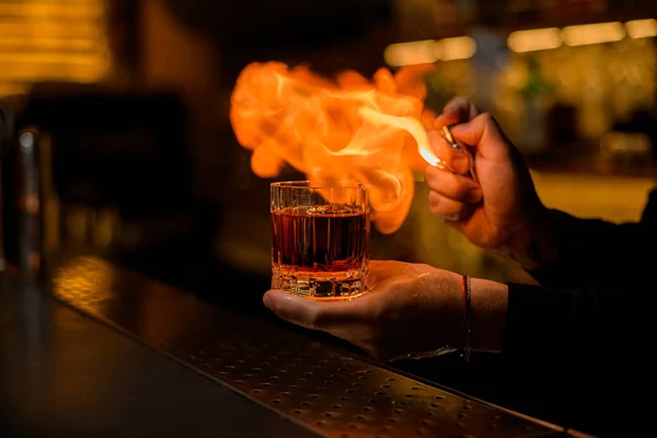 Hand of bartender hold old-fashioned glass with alcoholic drink and a piece of ice and make fire near it — Stock Photo, Image