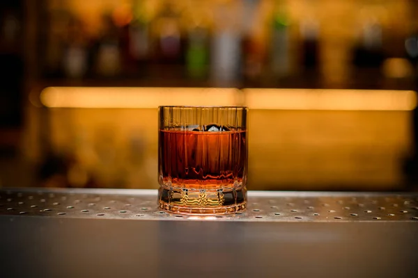 Front view of old-fashioned glass with alcoholic drink and piece of ice on blurred background — Stock Photo, Image