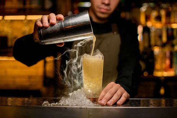 Mano del camarero sostiene agitador y vierte suavemente un cóctel humeante en un vaso con trozos de hielo en el mostrador de la barra — Foto de Stock