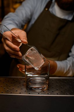 Erkeğin elindeki maşalarla şeffaf camların üzerinde duran büyük buz küpünün görüntüsü