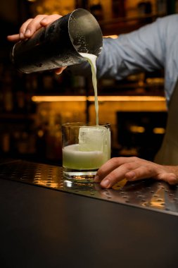 Barmenin eli titreticiyi tutar ve bardağa bir kokteyl döker.