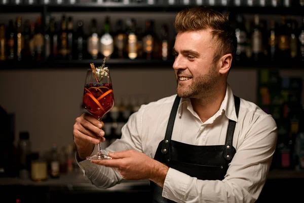 Sorridente barman masculino detém vidro com coquetel assinatura em suas mãos e olha para ele — Fotografia de Stock