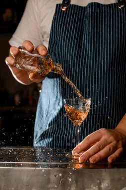 Barmen elinde küçük bir şişe tutuyor ve alkollü bir içkiyi bardağa dolduruyor.