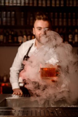 Barmen elinde alkollü bir içecekle bardağı tutuyor ve elini dumanın içinden uzatıyor.