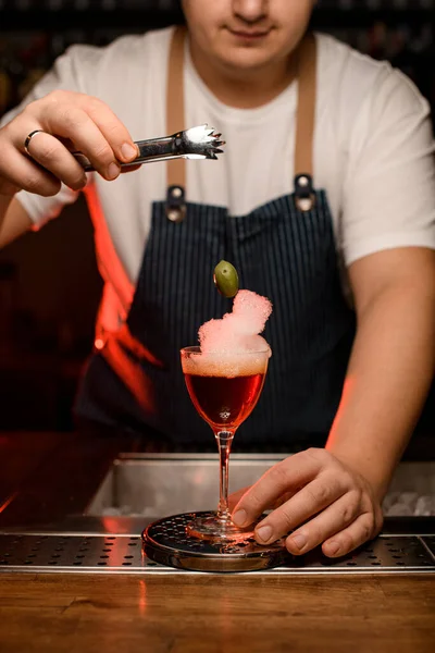 Bartender använder tång för att sätta en oliv i glas fylld med röd skummande alkoholhaltig dryck — Stockfoto