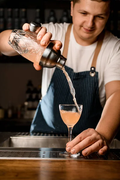 Bartender prende profissionalmente por duas mãos coqueteleira vítrea com coquetel alcoólico marrom frio e agita-lo . — Fotografia de Stock