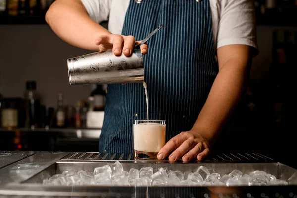 Bartender pours cocktail from shaker into glass standing on metal bar counter — Stock Photo, Image