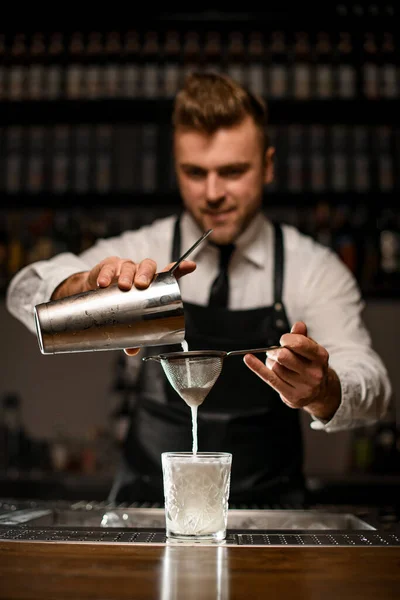 Barman filtra el cóctel blanco de la taza del agitador de acero en el vidrio pasado de moda a través del tamiz. —  Fotos de Stock