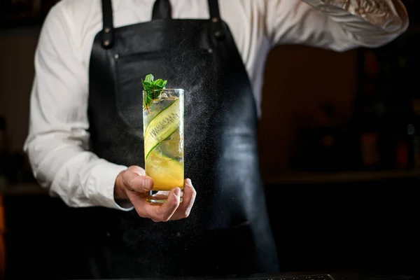 Hand des Barmannes hält und streut sanft auf Trinkglas mit kaltem Cocktail, der mit Minzzweigen und Gurkenstreifen verziert wird — Stockfoto