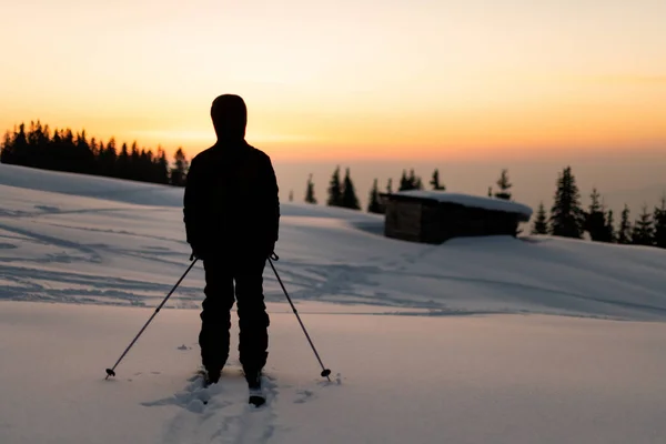 Bakifrån av människan med skidor och skidstavar som står i snön mot bakgrund av solnedgången himlen — Stockfoto