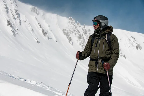 Męski narciarz w kurtce z goglami, hełmem narciarskim i kijami narciarskimi stoi na tle ośnieżonych gór — Zdjęcie stockowe