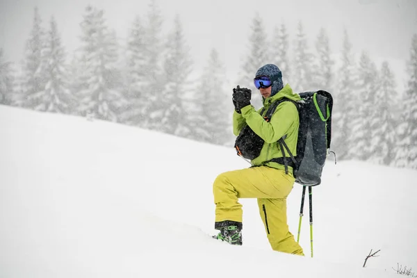 Vue latérale du skieur masculin en combinaison de ski verte et jaune avec sac à dos debout seul et prenant des photos de la zone enneigée — Photo