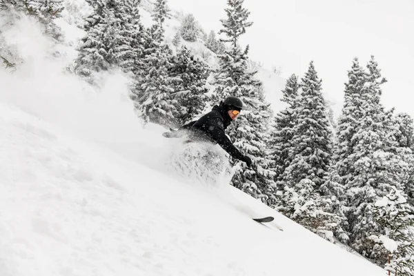 Narciarz w czarnym stroju narciarskim, hełmie i goglach zjeżdżający po pokrytych śniegiem stokach na nartach — Zdjęcie stockowe