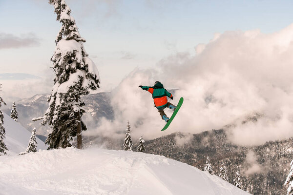 Snowboarder freerider jumping from snow-capped mountain slope on background of trees and cloudy sky