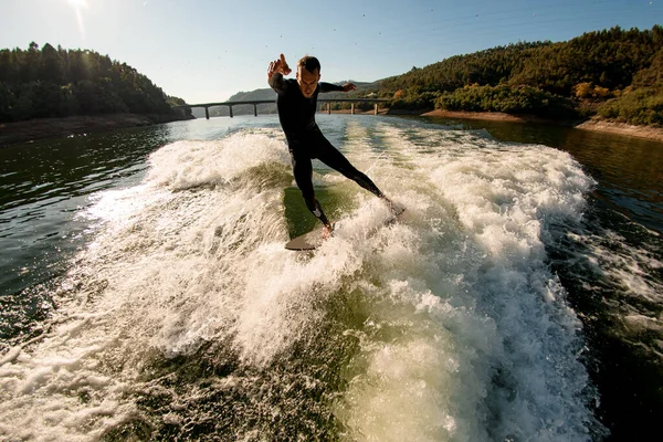 Man in black wetsuit balancing on splashing wave on a wakesurf — Stockfoto