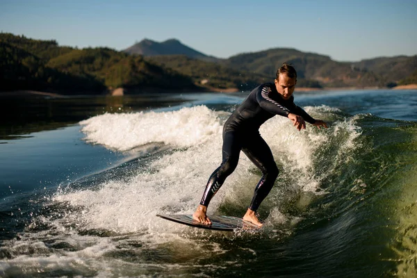 Attractive flexible man in wetsuit on wakesurf skilfully riding on splashing wave — Stockfoto