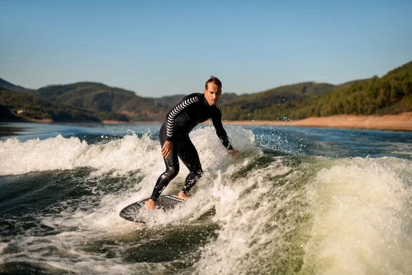 Man in black wetsuit rides on splashing river wave on a wakesurf — Stockfoto