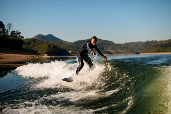 Flexible man in wetsuit on wakesurf masterfully riding on splashing river wave — Stockfoto