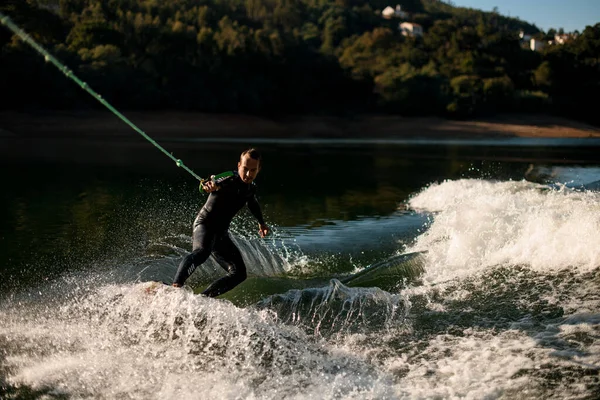 Sports man holds rope with handle and vigorously riding on wakesurf board on splashing water. — Stockfoto