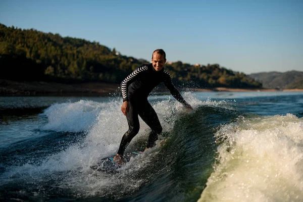 Cheerful man in wetsuit on wakesurf riding down the splashing wave — Stockfoto