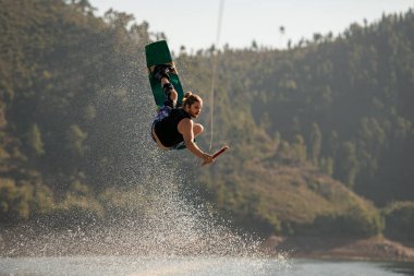 Aktif erkek sürücü halatı tutuyor ve wakeboard üzerinde aşırı atlıyor.