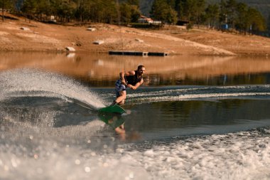 Atletik sakallı adam ipi tutuyor ve ustalıkla nehirde wakeboard sürüyor..
