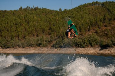 Çekici sakallı adamın harika bir manzarası var. Elinde ip var ve Wakeboard 'da nehrin üzerinden atlıyor.