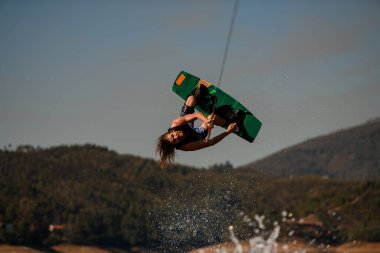 Aktif adam Wakeboardcu gökyüzüne ve tepelere karşı su sıçratan dalgalar üzerinde takla atıyor.