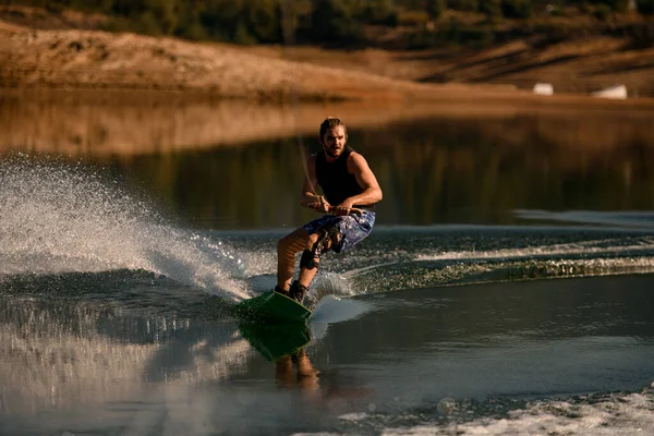 Atletico barbuto ragazzo tiene corda e rapidamente cavalcando wakeboard sul fiume. — Foto Stock