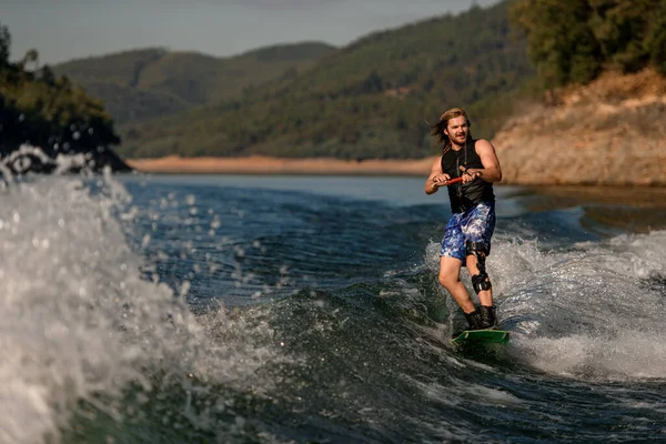 Attractive bearded guy holds rope and riding on the river wave on wakeboard — Fotografia de Stock