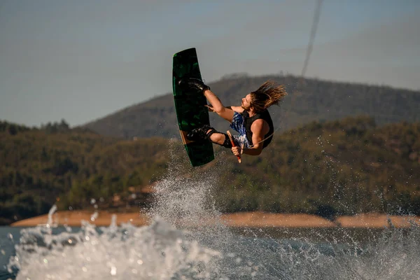 Wakeboarder hace acrobacias extremas saltando y voltea en wakeboard sobre salpicar ola contra el cielo y las colinas —  Fotos de Stock