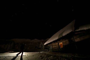 Kayak yapan bir adam ve kayak kazıklarıyla, alacakaranlıkta ahşap kulübeye doğru yürüyor.