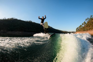 Aktif bir adam güzel bir manzara arka planında flama dalgasının üzerinden ustalıkla atlıyor. Yaz zamanı su sporları aktivitesi