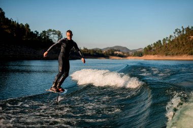Siyah dalgıç kıyafetli genç adam güzel bir manzara arka planında, bir folyo wakeboard üzerinde enerjik bir şekilde dengede duruyor..