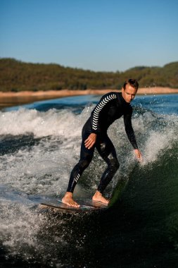 Siyah dalgıç kıyafetli aktif atletik adam dalgalanan nehir dalgası üzerinde wakesurf tahtasıyla ustaca ilerliyor.