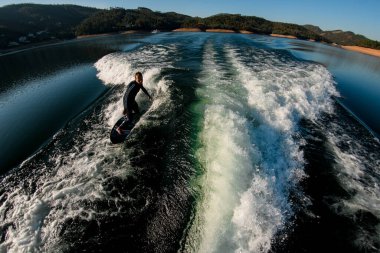 Siyah dalgıç kıyafetli genç bir sporcunun Wakesurf tahtasındaki bir dalgada dengede duruşu.